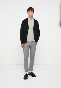 Čierny pletený kardigán over prúžkovanou košeľou s gombíkmi, šedé slim-fit nohavice a čierne mokasíny, doplnené bielymi ponožkami. Minimalistický dizajn.