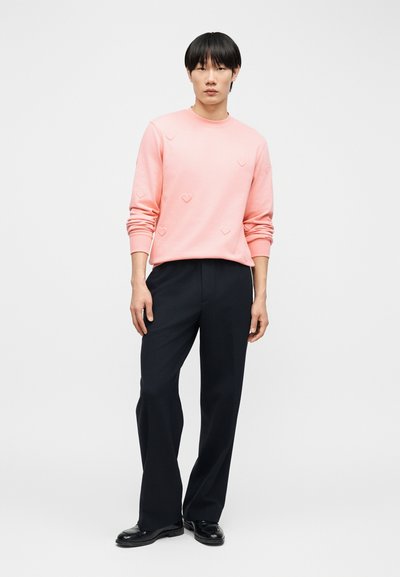 Νέος άνδρας που φοράει ανοιχτό ροζ μακρυμάνικο φούτερ με μικρά μοτίβα καρδιών, μαύρο παντελόνι με φαρδιά μπατζάκια και μαύρα παπούτσια, στέκεται όρθιος.