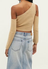 Asymmetrische beige gebreide top met lange mouwen; heeft een open schouderontwerp. Gecombineerd met een lichtblauwe denim rok, toont de achterkant.