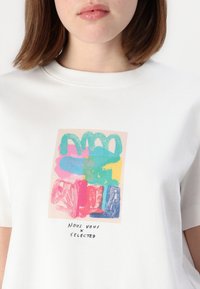 T-shirt en coton blanc avec un design abstrait coloré comprenant des coups de pinceau roses, bleus, verts et jaunes. Le texte en dessous indique "NOUS VOUS x SELECTED."