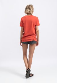 Rode katoenen t-shirt met korte mouwen, achterkant gezien, gedragen met een donkere short en zwarte sandalen, tegen een witte achtergrond.