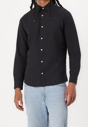 Personne portant une chemise noire boutonnée à manches longues et un jean bleu clair, debout avec les bras détendus le long du corps.