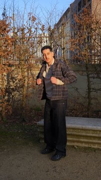 Άνδρας που προσαρμόζει το καρό σακάκι του ενώ στέκεται σε χωμάτινο μονοπάτι δίπλα σε λιθόκτιστο παγκάκι, με γυμνά δέντρα και κτίρια στο βάθος.