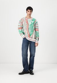 Pull en tricot avec un motif mélangé de vert et de rouge, présentant divers designs abstraits. Accompagné de jean bleu et de chaussures noires.