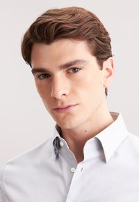 Giovane uomo con capelli corti e ondulati di colore marrone, indossa una camicia button-up grigia chiara con un motivo, e guarda direttamente la macchina fotografica.