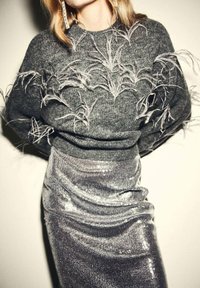 Pull en maille gris avec des embellissements en textures ressemblant à des plumes. Jupe en sequins argent métallisé avec des détails froncés, créant un effet superposé.