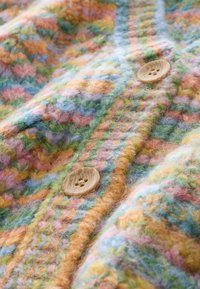 Cardigan en laine douce avec motif zigzag pastel multicolore et deux boutons marron clair à quatre trous sur un tissu tricoté texturé.