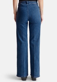 Personne portant un jean en denim bleu taille haute et des chaussures à talons noires, se tenant de dos sur un fond blanc uni.