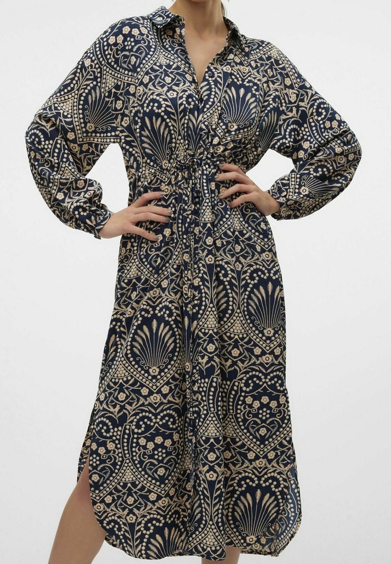 Femme portant une robe bleu marine à manches longues avec des motifs floraux et géométriques beiges, taille cintrée, col et fente latérale, mains sur les hanches.