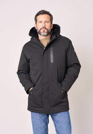 Homme barbu portant un manteau d'hiver noir à capuche et un jean bleu, debout les mains dans les poches du manteau devant un fond uni.