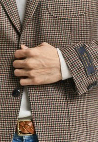 Person wearing houndstooth blazer with GANT label on sleeve, white sweater, brown braided belt, and blue jeans, hand adjusting blazer.