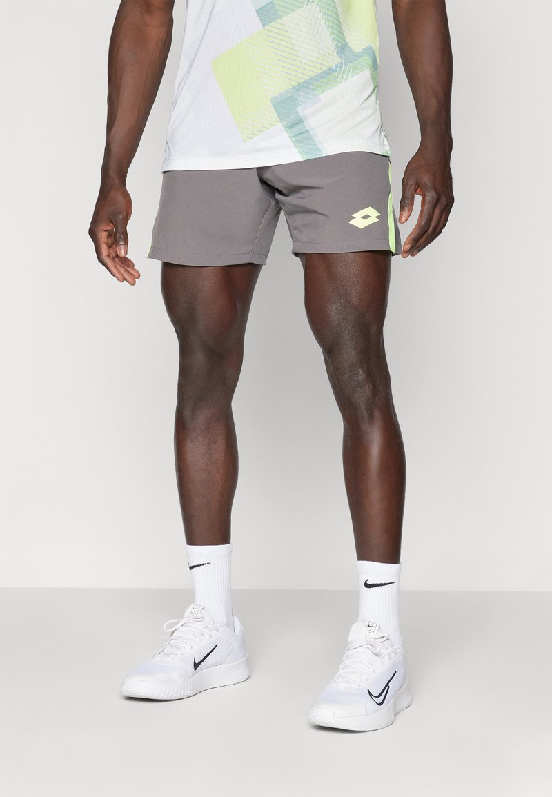 Pantalones cortos deportivos grises con un acento amarillo, combinados con zapatillas y calcetines blancos de Nike. La parte superior presenta patrones geométricos en colores claros.