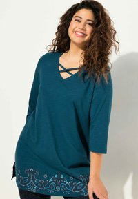 Teal langærmet skjorte med V-hals og dekorative krydsslynger. Har indviklede blomster mønstre i blå og mørke nuancer ved kanten.