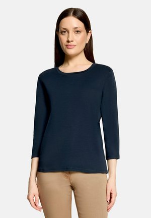 Femme aux cheveux bruns lisses portant une chemise bleu marine à manches longues et un pantalon beige, debout sur un fond uni.