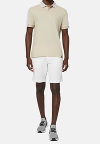 Polo beige avec un col blanc contrastant et des manches courtes, associé à un short blanc. Le mannequin porte des chaussures de sport grises.