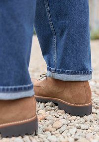 Botine din suede maro, cu o talpă de tracțiune închisă la culoare, asociate cu blugi blue denim rulați, așezate pe un teren de pietriș.