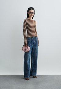 Striped long-sleeve top in brown and white, paired with high-waisted blue denim wide-leg jeans. Model holds a pink, gathered handbag.