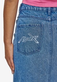 Blå denimskjørt med middels høy midje, med baklomme og hvit grafisk logo design. Stoffet har en teksturert finish.