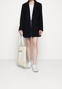 Cappotto nero su misura con un bottone singolo, due tasche laterali, indossato sopra una maglietta bianca e pantaloni corti di jeans, abbinato a sneakers bianche e a una borsa tote.