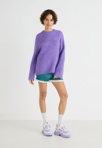 Lila bolyhos pulóver, teal színű, csipkés szegélyű sort, és tömör lila-fehér sportcipő. A modell egyszínű háttér előtt áll.