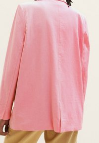 Svetle růžový blazer vyrobený z texturované látky, s klopovým límcem a bočními rozparky. Design je volný s dlouhými rukávy.