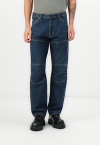 Άνδρας που φοράει σκούρα μπλε τζιν παντελόνια με μπαλώματα στα γόνατα και μαύρα χοντρά παπούτσια, στέκεται μπροστά σε λευκό φόντο.