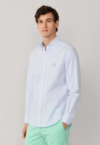 Chemise à rayures bleues et blanches, à boutons, avec un col, des manches longues et un petit logo brodé sur la poitrine. Texture de tissu lisse.