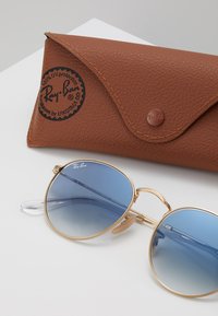 Occhiali da sole Ray-Ban con lenti rotonde a sfumatura blu chiaro, montatura in metallo dorato e una custodia in pelle marrone con logo impresso.
