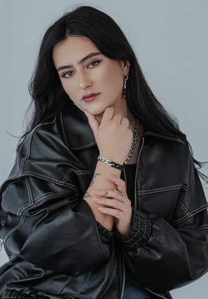 Black leather jacket with white stitching, worn by a person. Features a silver chain bracelet and multiple rings on hands. Smooth texture.