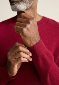 Karmesinrot gestrickter Pullover mit gerippten Handgelenksbündchen. Nahaufnahme von Händen, die die strukturierte Haut und die Strickdetails zeigen. Einfaches, elegantes Design.