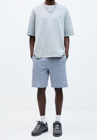 Un sweatshirt oversize gris clair à manches courtes, associé à un short bleu avec cordon de serrage. Des baskets noires avec des détails texturés complètent la tenue.