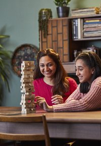 Jenga game made of light wood blocks, stacked vertically, with two players in colorful knit sweaters, engaged around a wooden table.