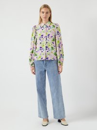 Blusa floral com padrões roxos e verdes, design de botões, mangas longas, combinada com jeans de cintura alta azul claro e sapatos de salto bicolor.