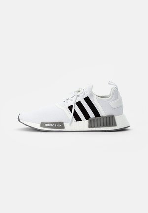 Baskets blanches Adidas avec des bandes noires, des accents gris sur la semelle et des lacets blancs, vues de profil sur un fond uni.