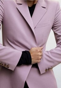 Blazer violet lavande ajusté avec des revers structurés, une poche poitrine simple et des poignets boutonnés. Le tissu semble lisse et cintré.
