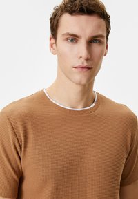 Brun struktureret t-shirt med rund hals, prydet med hvid kant. Stoffet har et vaffelmønster og korte ærmer.