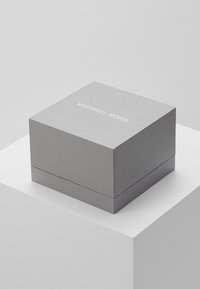 Caja de regalo cuadrada gris con una superficie texturizada, que presenta el texto en relieve "MICHAEL KORS" en blanco en la tapa, ligeramente abierta.
