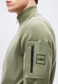 Άνδρας που φοράει πλεκτό πουλόβερ ελιάς με ψηλό λαιμό, με το λογότυπο BOSS και κάθετρη τσέπη με φερμουάρ στο αριστερό πάνω μανίκι.
