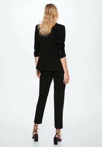 Black tailored blazer with structured shoulders and long sleeves, paired with slim-fit black trousers and heeled sandals. Smooth fabric.