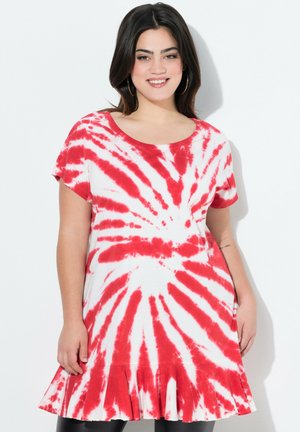 Mujer sonriente con cabello oscuro, lleva pendientes de aro y un vestido tie-dye rojo y blanco con mangas cortas y un dobladillo con volantes.