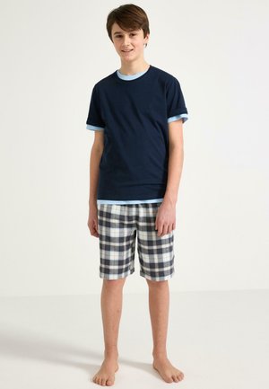 Teenagedreng står barfodet iført en mørkeblå t-shirt over en lyseblå skjorte og ternede shorts mod en ensfarvet hvid baggrund.