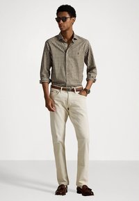 Καρό πουκάμισο με κουμπιά σε χρώματα καφέ και μπεζ, ανοιχτό μπεζ παντελόνι chino, καφέ ζώνη, γυαλιά ηλίου, στέκεται με τα χέρια στις τσέπες. Υφή με κηλίδες στο πουκάμισο.