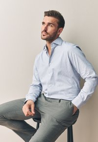 Uomo con camicia azzurra a quadri e pantaloni grigi seduto su uno sgabello, che guarda pensieroso verso l'alto su uno sfondo semplice.