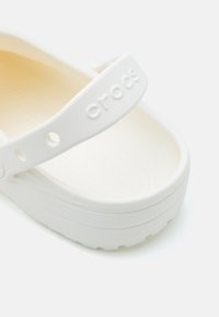 Sandal din cauciuc alb cu o curea lată ce prezintă logo-ul "Crocs". Talpă lată cu un interior texturat și margini rotunjite. Design ușor.