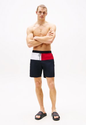 Men's swim shorts featuring a color block design in navy, red, and white with an elastic waistband and drawstring. Worn with black flip-flops.