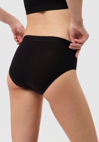 Eine Frau trägt schwarze hochgeschneiderte Unterwäsche und ein schwarzes Crop-Top, während sie den Bund anpasst, von hinten vor einem schlichten Hintergrund gezeigt.