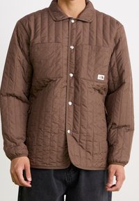 Mężczyzna w brązowej pikowanej kurtce z zatrzaskami, kieszeniami na piersi i elastycznymi mankietami, w połączeniu z ciemnymi spodniami, na jednolitym tle.