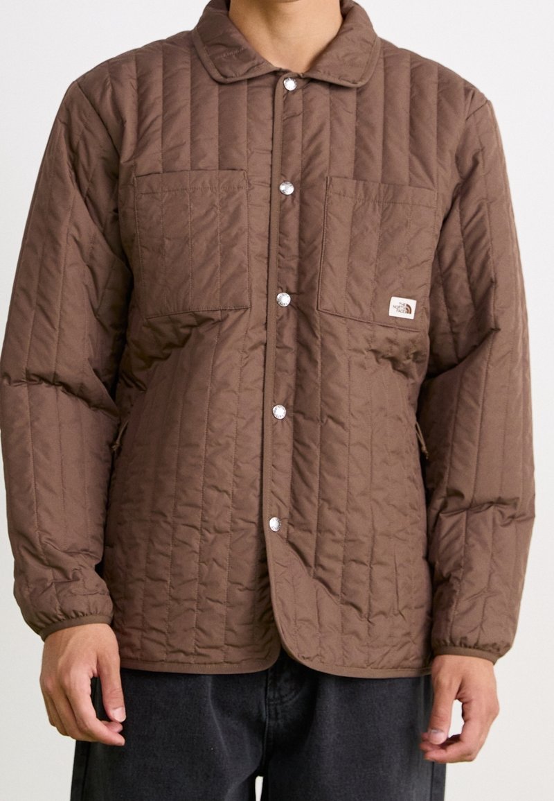 Mężczyzna w brązowej pikowanej kurtce z zatrzaskami, kieszeniami na piersi i elastycznymi mankietami, w połączeniu z ciemnymi spodniami, na jednolitym tle.