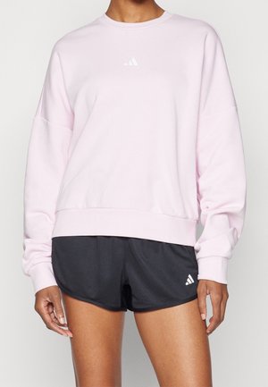 Persoon draagt een lichtroze Adidas sweatshirt en zwarte Adidas sportshort, staand tegen een neutrale effen achtergrond.
