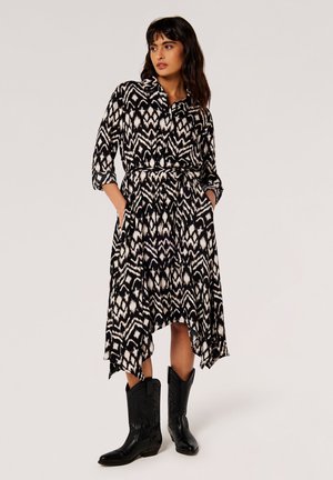 Kvind i en sort-hvid mønstret kjole med en lommetørklæde-saum, ¾ ærmer, hænderne i lommerne, iført sorte cowboystøvler.